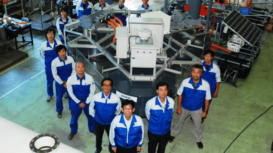 水流発生装置ジェットストリーマーを囲んで立つ、青と白の作業着を着用した技術者・スタッフの集合写真