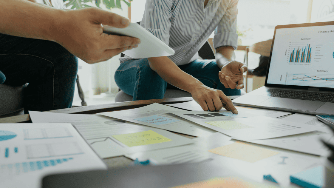 会議テーブルに並べた企画書や資料を囲み、グラフを表示したノートPCを見ながら商品企画の打ち合わせをする担当者たち