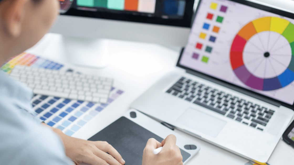 カラーチャートや色相環を表示したモニターとパソコンを前に、ペンタブレットでデザイン企画を行う担当者の手元